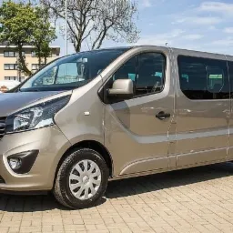 Beżowy Opel Vivaro, widok z przodu i boku, zaparkowany na brukowanym placu, budynek w tle.