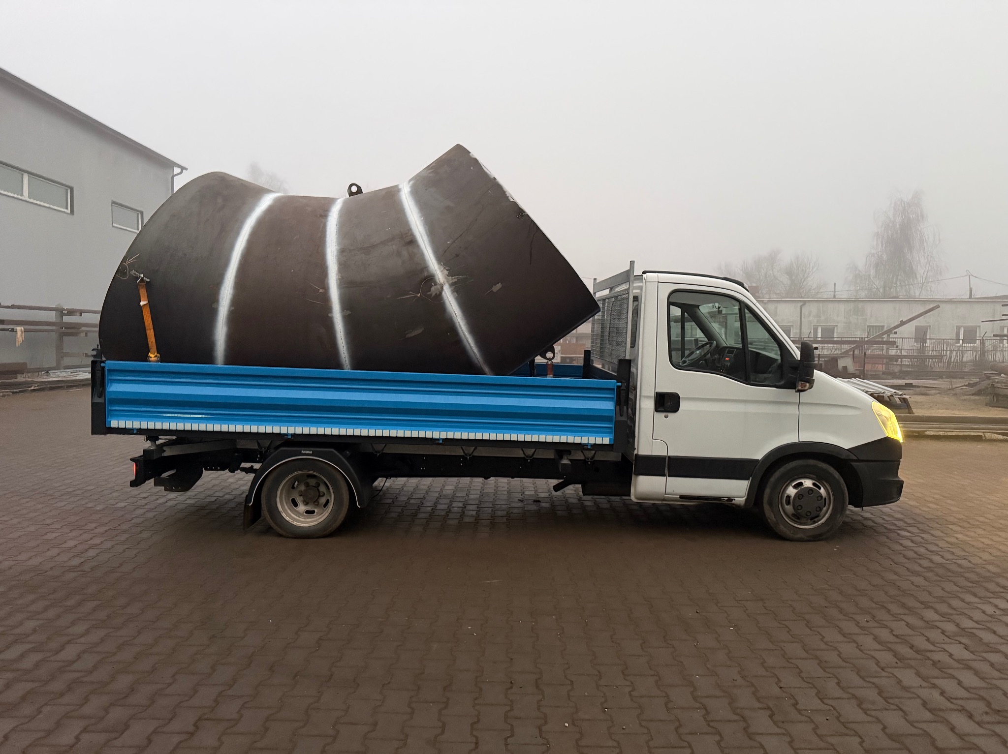 Biały bus z niebieską skrzynią transportuje duży, metalowy element z białymi pasami. Mglista pogoda, budynek w tle. Widok z boku na kostce brukowej.