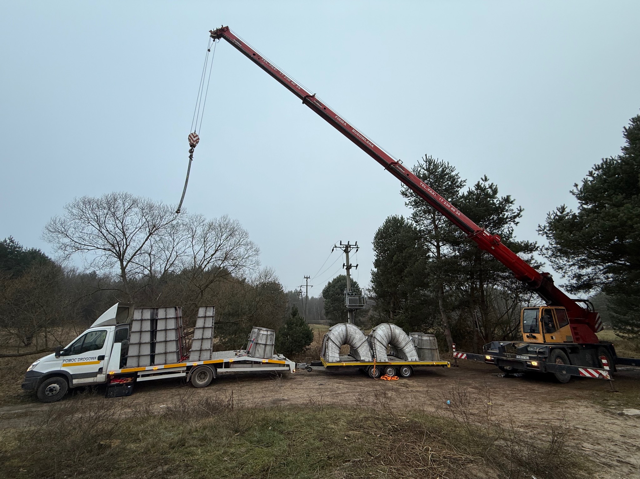 Ciężarówka pomocy drogowej z przyczepą przewozi elementy konstrukcyjne pod nadzorem dźwigu teleskopowego na tle leśnego krajobrazu w pochmurny dzień.
