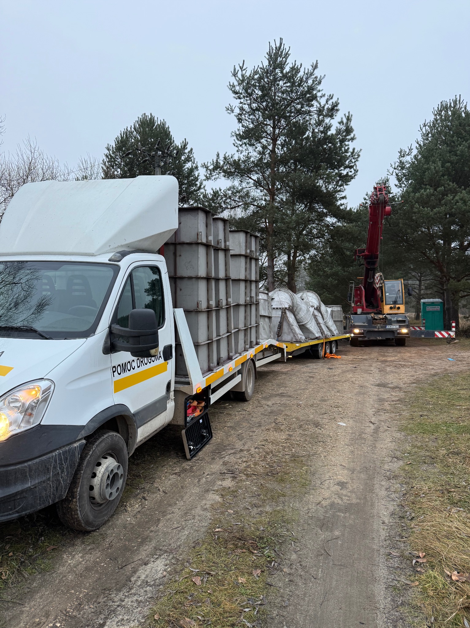 Laweta z napisem 'Pomoc Drogowa' przewozi metalowe elementy konstrukcyjne. W tle żuraw i przenośna toaleta na leśnej drodze.