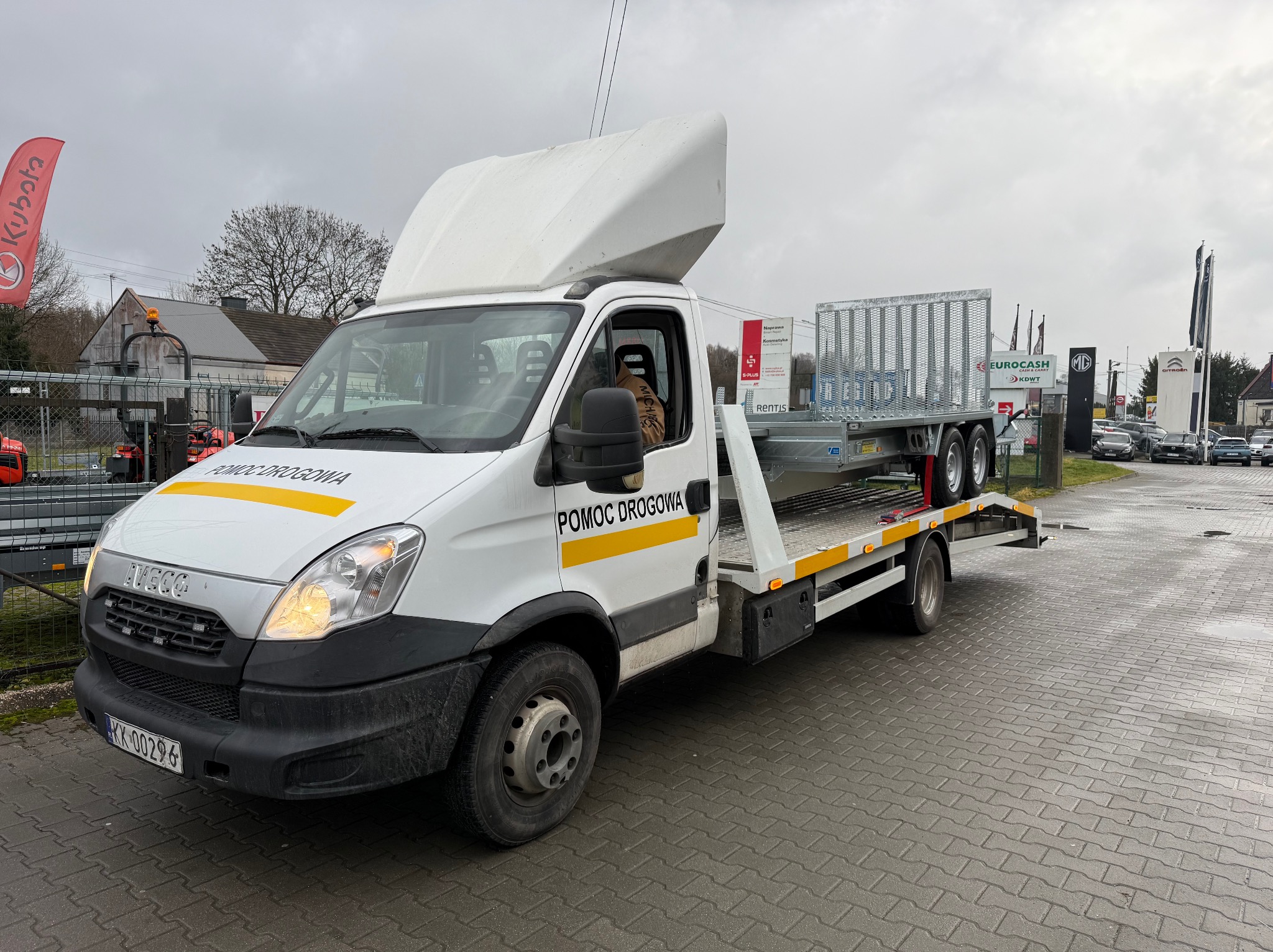 Biała laweta Iveco z żółtymi pasami i napisem 'Pomoc Drogowa' na parkingu, z przyczepą na platformie. W tle budynki i reklamy salonów samochodowych.