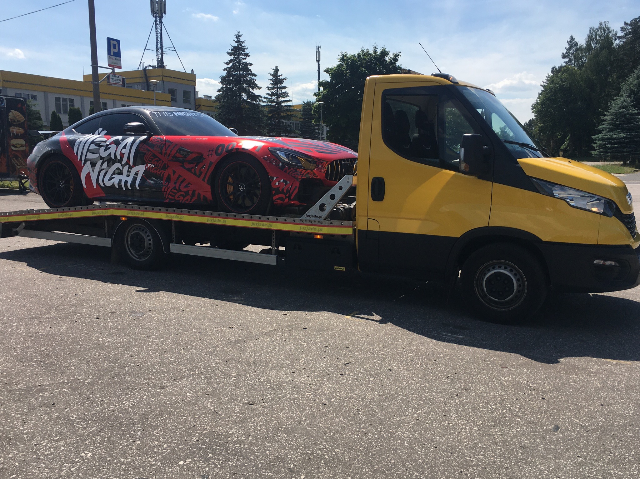 Żółta laweta z czarnymi elementami przewozi czerwono-czarny sportowy samochód marki Mercedes z napisem 'MEGA NIGHT' na boku, zaparkowana na szarym asfalcie, w tle budynek i drzewa.