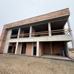 HOCH BAU DAWID ŚLUSARCZYK - Surowy stan dwukondygnacyjnego budynku z betonu i cegły, widok z zewnątrz na plac budowy w pochmurny dzień. Widoczne niezabezpieczone otwory okienne i drzwiowe.