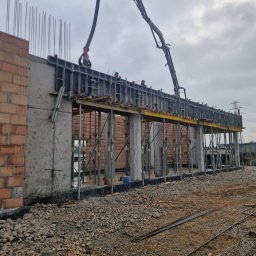 HOCH BAU DAWID ŚLUSARCZYK - Wylewanie betonu na strop budynku w trakcie budowy. Widoczne szalunki, zbrojenia i pracownicy w kaskach. Na pierwszym planie gruz i elementy zbrojeniowe.