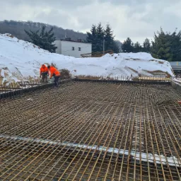 Zbrojenie płyty fundamentowej z widocznymi pracownikami w pomarańczowych kombinezonach, śnieżny nasyp ziemi w tle, pochmurne niebo.