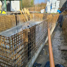 Zbrojenie fundamentu zalane betonem, widoczny wykop z rurą kanalizacyjną, w tle materiały budowlane zabezpieczone folią.