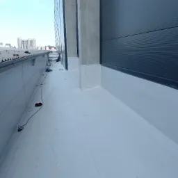 Płaski dach przemysłowy pokryty białą membraną hydroizolacyjną, widoczne obróbki blacharskie przy ścianie budynku, w tle instalacje przemysłowe.