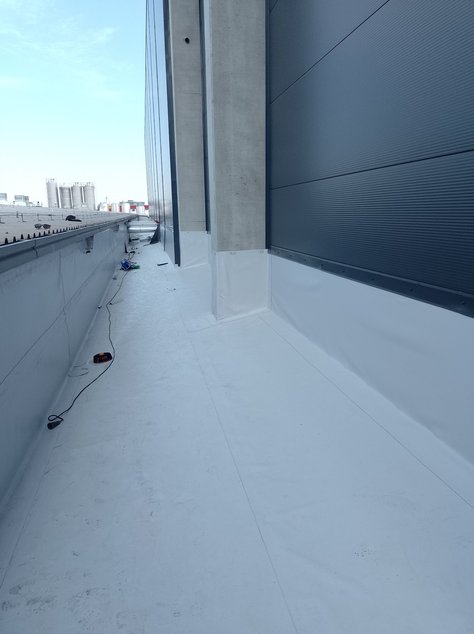Płaski dach przemysłowy pokryty białą membraną hydroizolacyjną, widoczne obróbki blacharskie przy ścianie budynku, w tle instalacje przemysłowe.