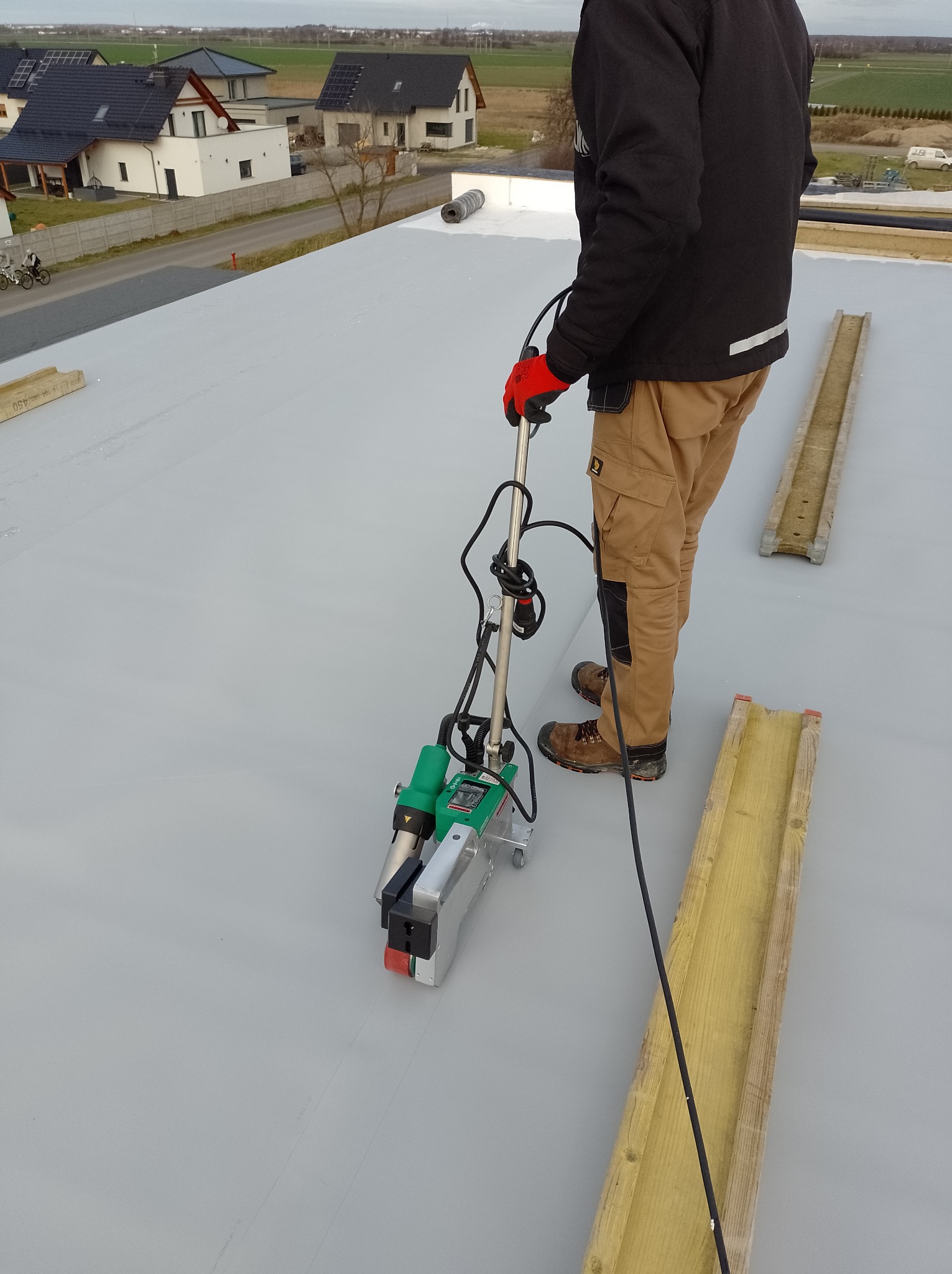 Pracownik w kombinezonie łączy arkusze szarej membrany dachowej za pomocą zgrzewarki na dachu nowego domu jednorodzinnego, widoczne drewniane prowadnice.