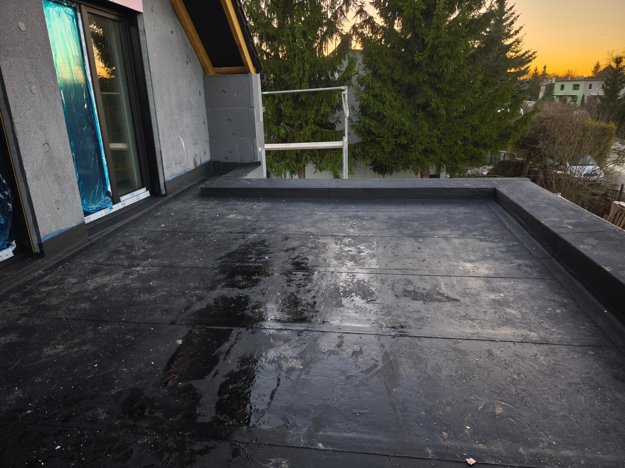 Czarna, płaska powierzchnia dachu pokryta papą, z widocznym oknem dachowym zabezpieczonym folią i fragmentem rusztowania na tle zielonych drzew i zachodzącego słońca.