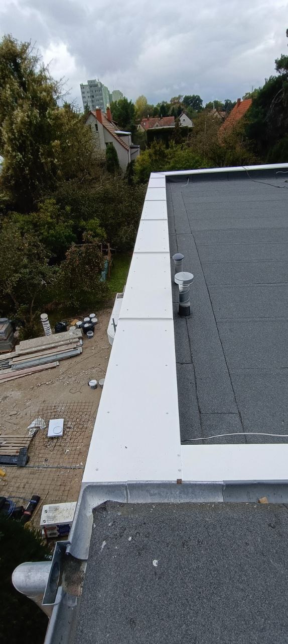 Szczegółowe ujęcie płaskiego dachu pokrytego papą, z widocznymi kominkami wentylacyjnymi i obróbkami blacharskimi w kolorze białym. W tle zieleń drzew i zabudowa miejska.