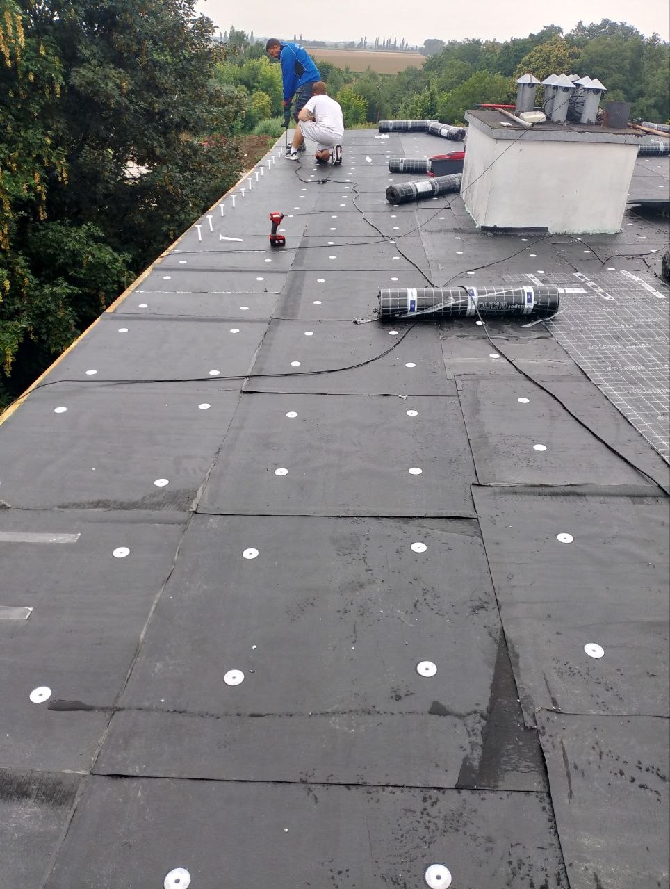 Dach pokryty czarną papą z białymi mocowaniami, dwóch dekarzy w trakcie pracy, widoczne rolki papy i narzędzia. Ujęcie z góry, krajobraz w tle.