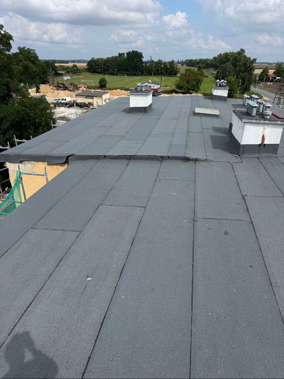 Szary, płaski dach pokryty papą termozgrzewalną, z widocznymi kominami i elementami wentylacji. W tle zieleń drzew i błękitne niebo z chmurami.