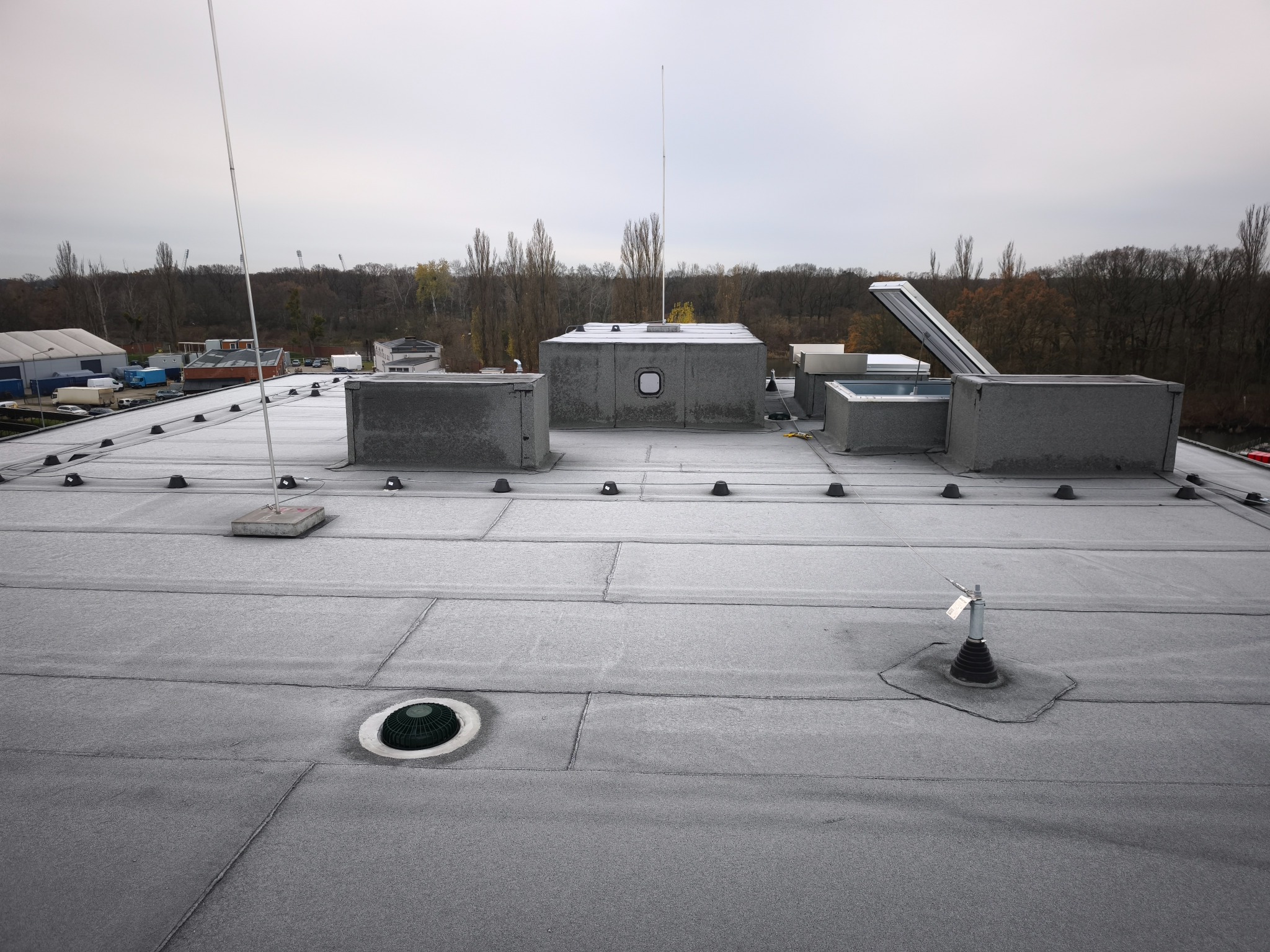 Szary dach płaski z widocznymi elementami wentylacyjnymi, odgromnikami i świetlikami dachowymi. W tle drzewa i budynki. Widok z góry, pochmurne niebo.