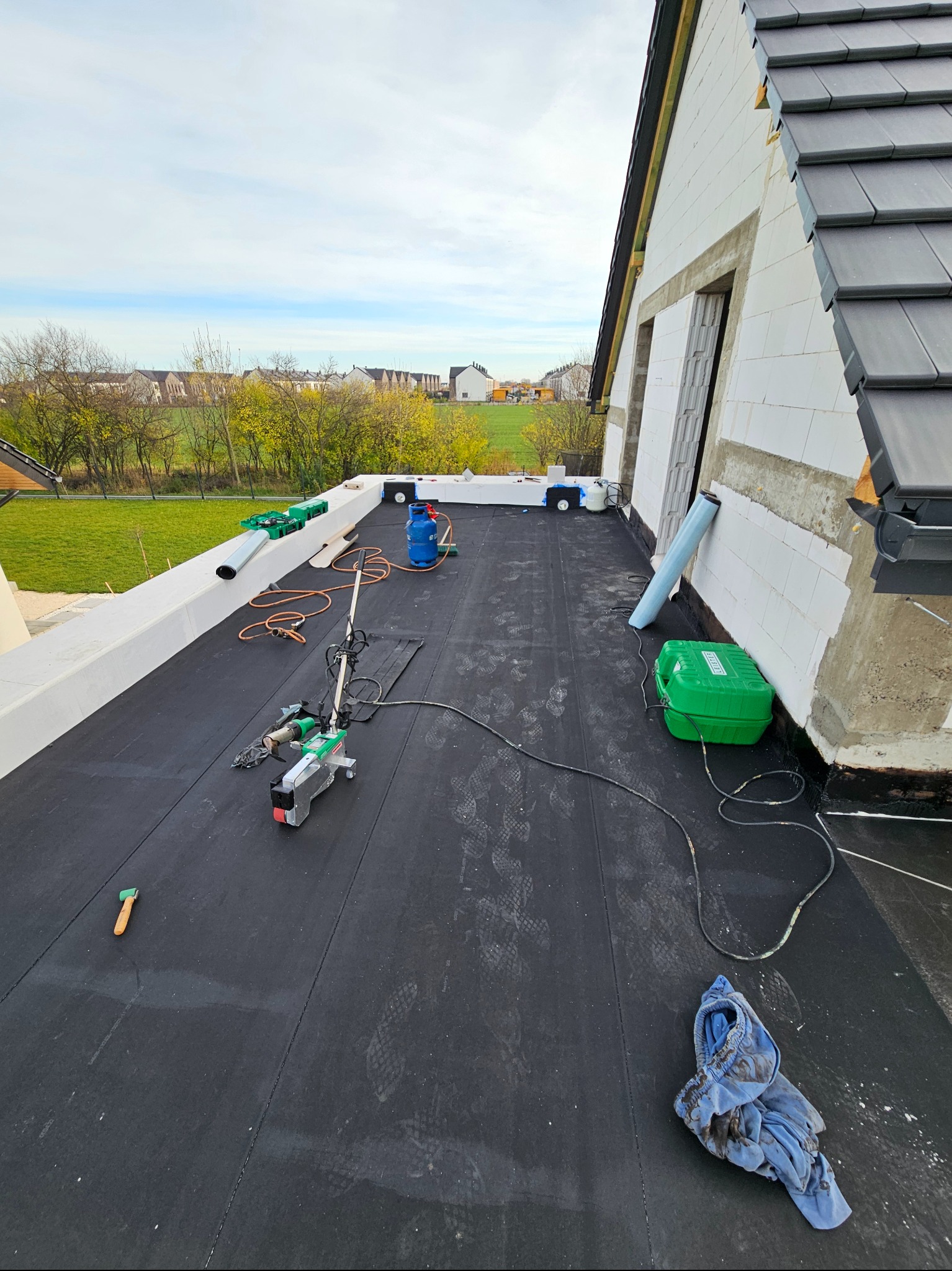 Czarna papa termozgrzewalna na dachu, narzędzia dekarskie, butla gazowa, widok na okolicę. Prace dekarskie w toku, z bliska, zarys budynku w tle.