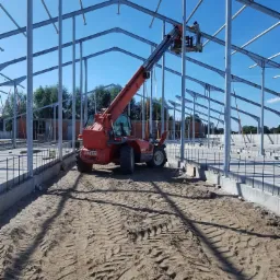Montaż stalowej konstrukcji hali z użyciem podnośnika teleskopowego Manitou, widoczne ślady opon na piaszczystym podłożu i zbrojenie fundamentów.