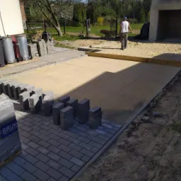 Ułożona część nawierzchni z kostki brukowej, przygotowany piasek, paleta z kostką w folii z widocznym logo producenta BRUK, pracownicy w tle.