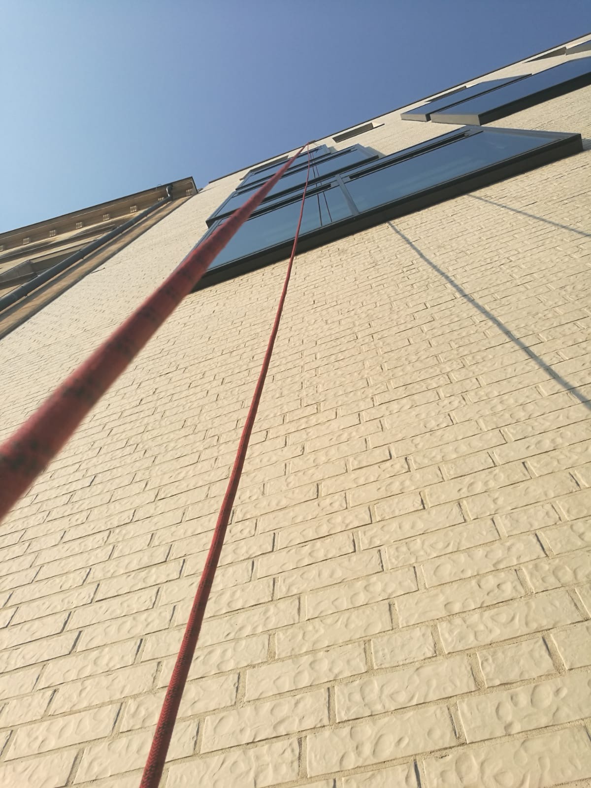 Widok z dołu na ścianę budynku z beżowej cegły z oknami i liną alpinistyczną, sugerujący prace na wysokościach.