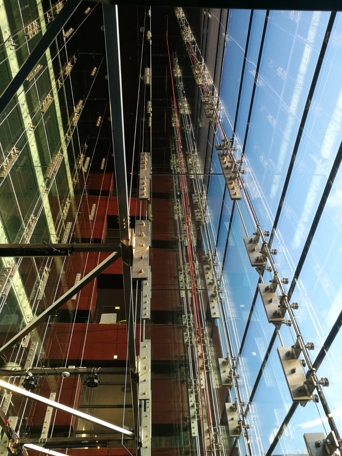 Szklana fasada budynku biurowego z widocznymi elementami mocującymi i systemem lin stalowych, odbijająca błękitne niebo.