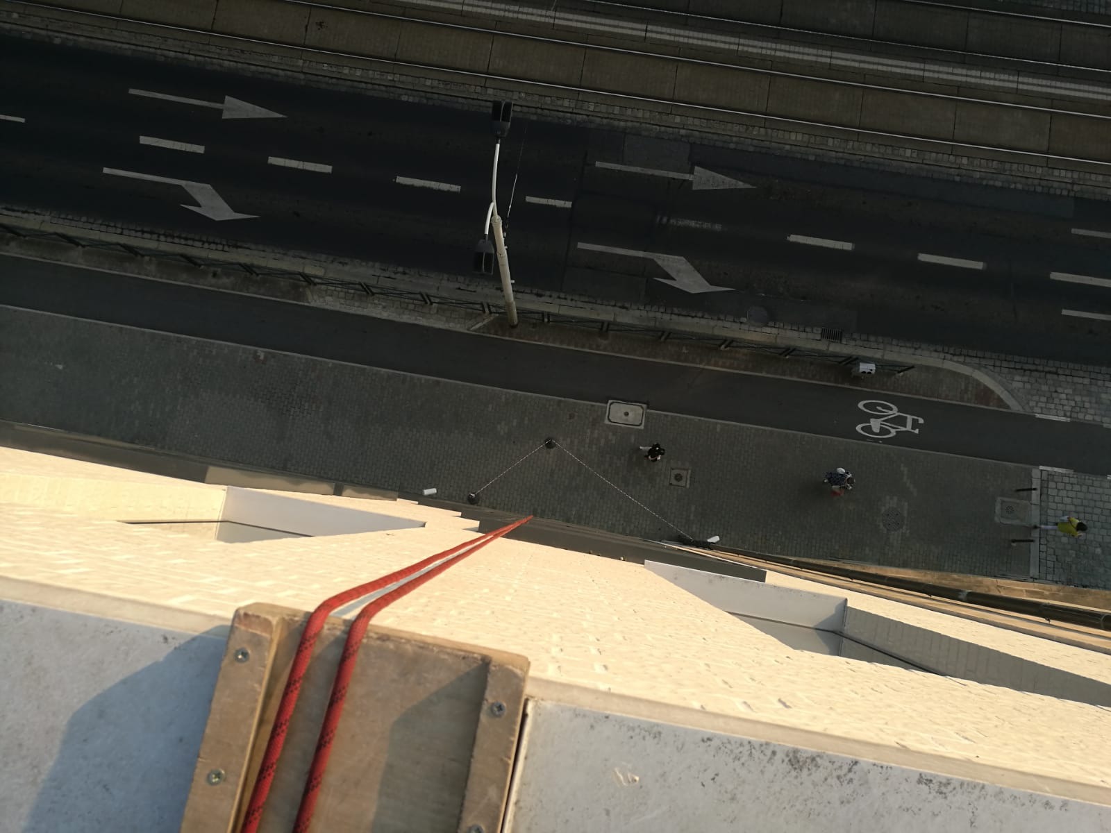 Widok z góry na ścianę budynku z zamocowanymi linami asekuracyjnymi, na chodniku poniżej widoczni pracownicy w trakcie wykonywania prac wysokościowych.