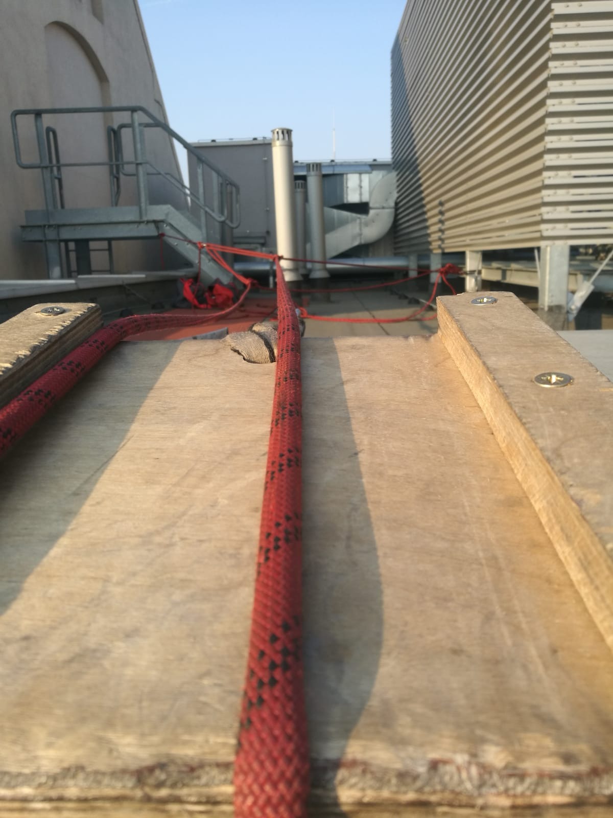 Czerwona lina zabezpieczająca rozciągnięta na drewnianej desce na dachu budynku z widocznymi elementami instalacji wentylacyjnej i schodami z metalową barierką.