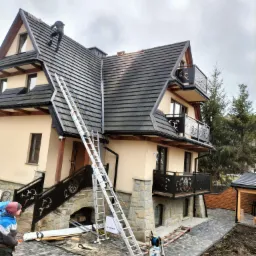 Wysoki dom w stylu zakopiańskim z kremową elewacją, kamienną podmurówką i ciemną dachówką. Na dachu pracuje dekarz, a obok stoi długa, rozłożona drabina. Widoczny fragment chodnika z kostki...