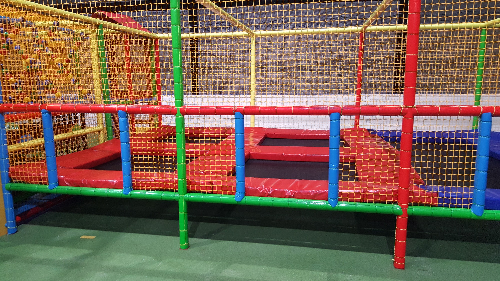 Wielopoziomowa konstrukcja zabawowa z siatką, trampolinami, kolorowymi rurami (czerwone, zielone, niebieskie, żółte) i basenem z kulkami w tle.