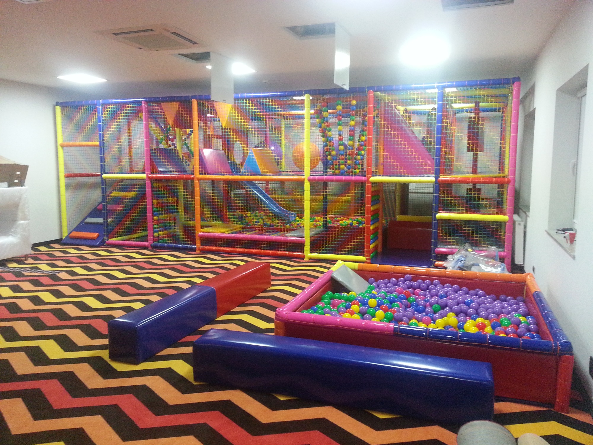 Wielopoziomowa konstrukcja do zabaw wewnątrz budynku z siatkami zabezpieczającymi, zjeżdżalniami i basenem z kolorowymi piłeczkami, uzupełniona o miękkie, podłużne siedziska w kolorach czerwonym...