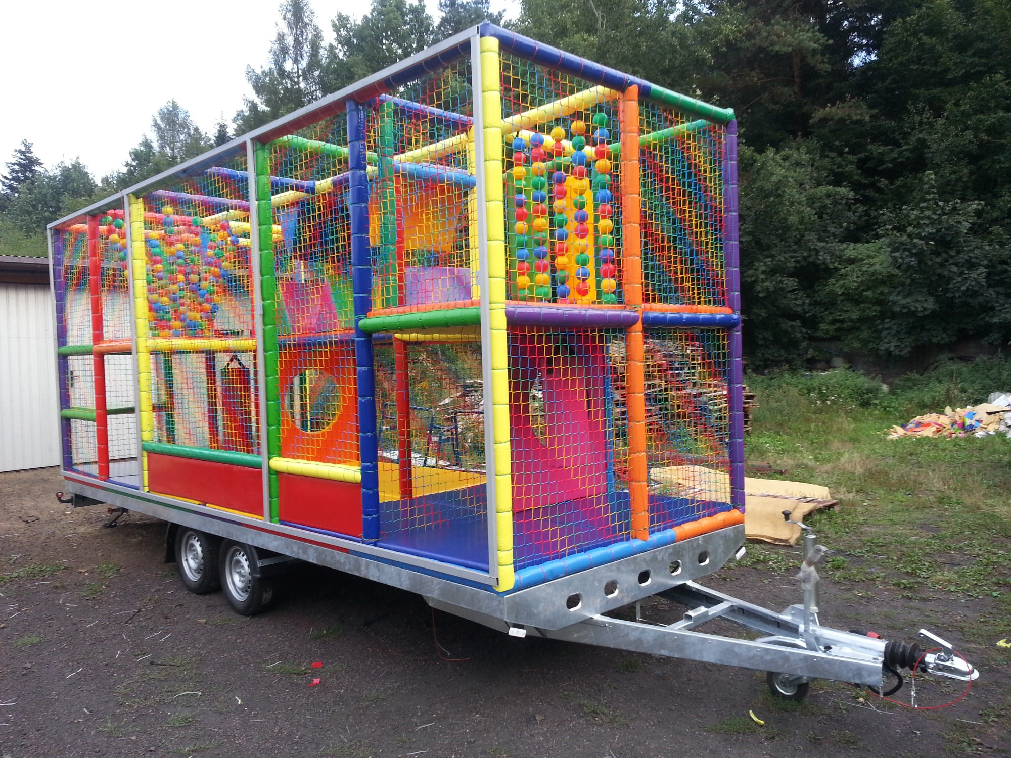 Przenośny, kolorowy plac zabaw na przyczepie, z siatką zabezpieczającą i elementami do wspinaczki, wypełniony kolorowymi piłeczkami.