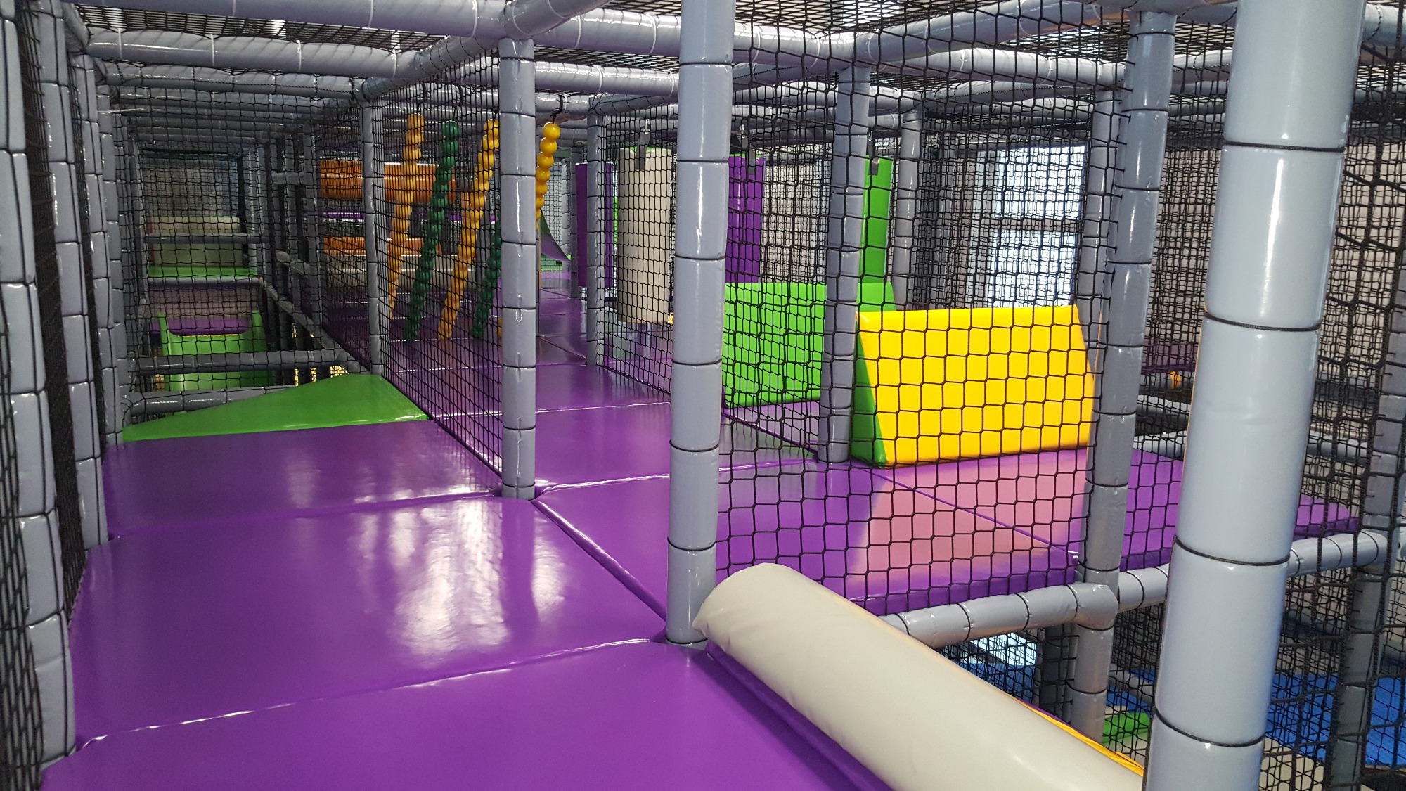 Wielopoziomowa konstrukcja wewnętrzna krytej sali zabaw z miękkimi elementami w kolorach fioletowym, zielonym i żółtym, zabezpieczona siatką, zrealizowana w Mykanowie.