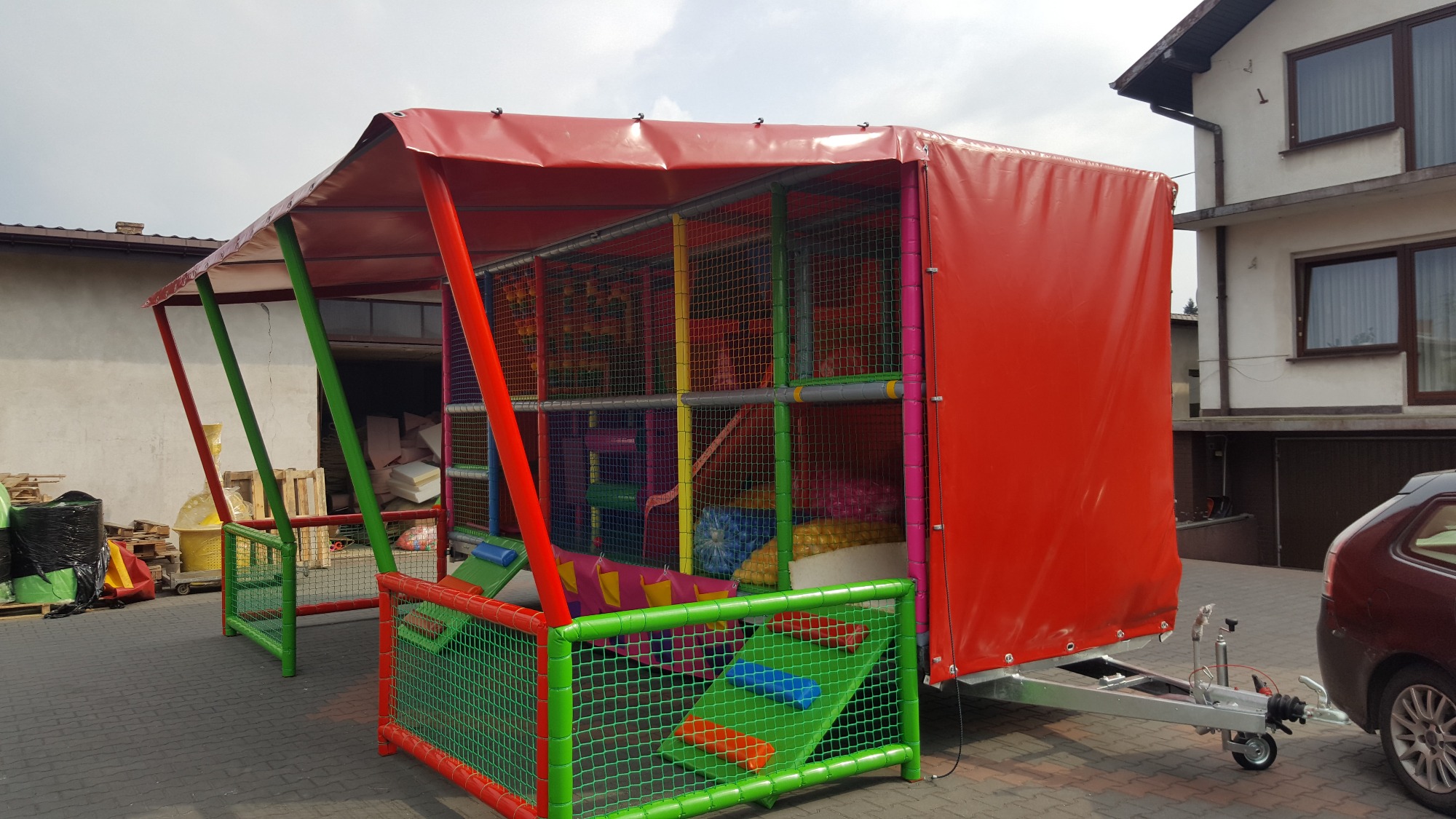 Przenośny, kolorowy plac zabaw na przyczepie, z zadaszeniem i siatkowymi ścianami, widoczny na tle budynków.