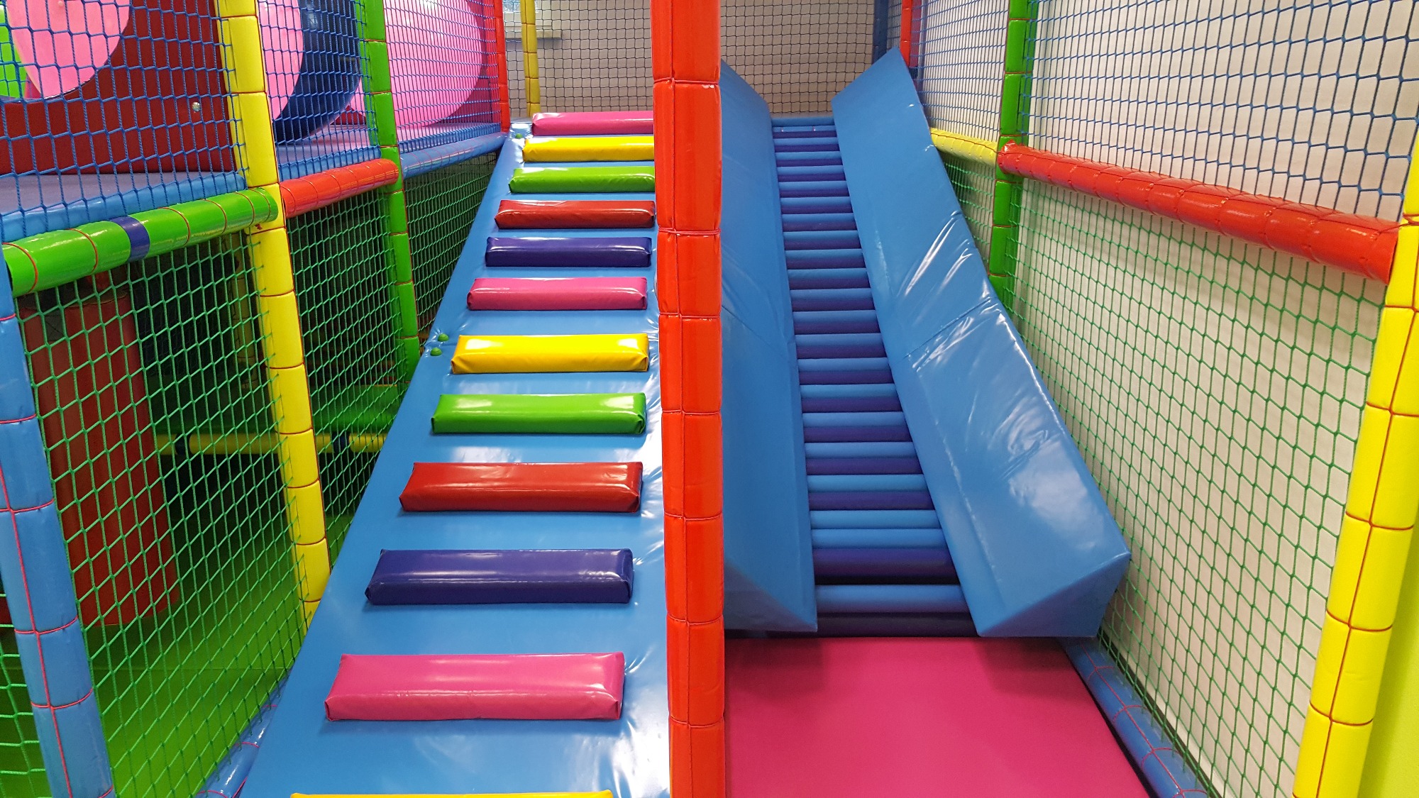 Wnętrze sali zabaw z kolorowymi schodkami i zjeżdżalnią, zabezpieczone siatką.
