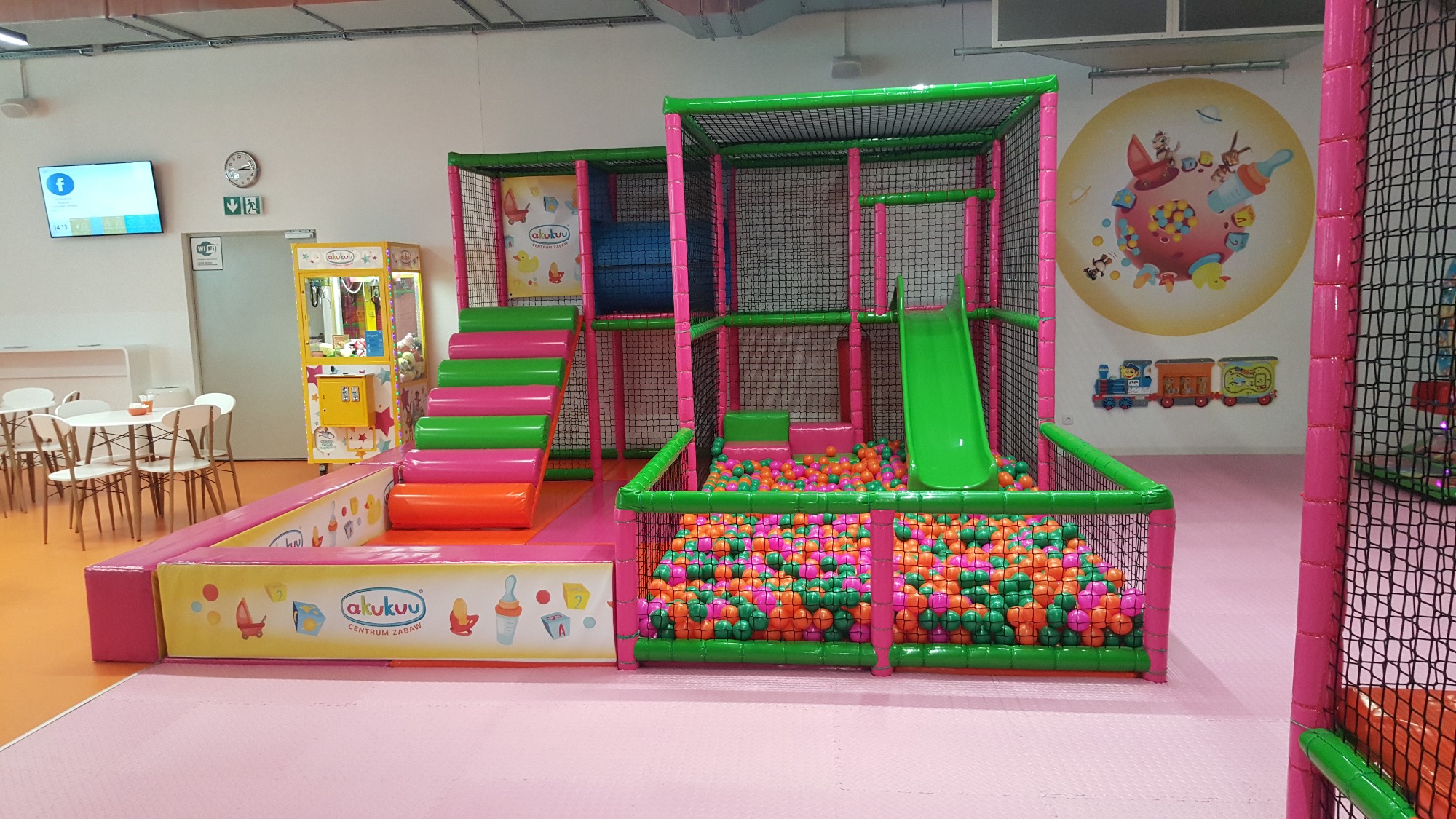 Wielopoziomowa konstrukcja zabawowa z basenem z kulkami, schodami i zjeżdżalnią w jasnych kolorach, z logo 'akukuu' na obudowie, w tle automat z zabawkami i stoliki z krzesłami.