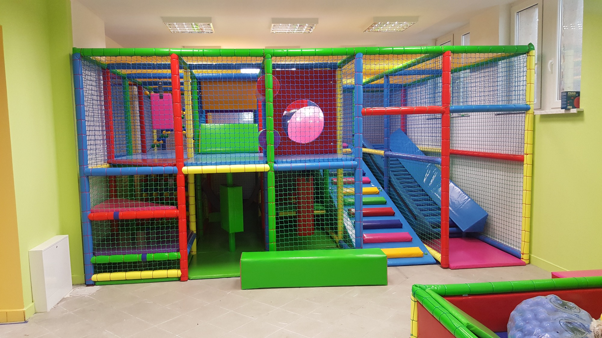 Wielopoziomowa konstrukcja zabawowa z siatkami ochronnymi, schodami i zjeżdżalnią, utrzymana w jaskrawych kolorach, umieszczona w jasnym pomieszczeniu z oknem.