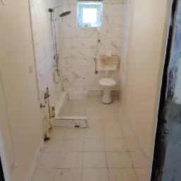 Łazienka w trakcie prac wykończeniowych: białe płytki imitujące marmur na ścianach, częściowo ułożone, widoczny brodzik prysznicowy z zamontowaną armaturą, toaleta z założonym ręcznikiem, podłoga...