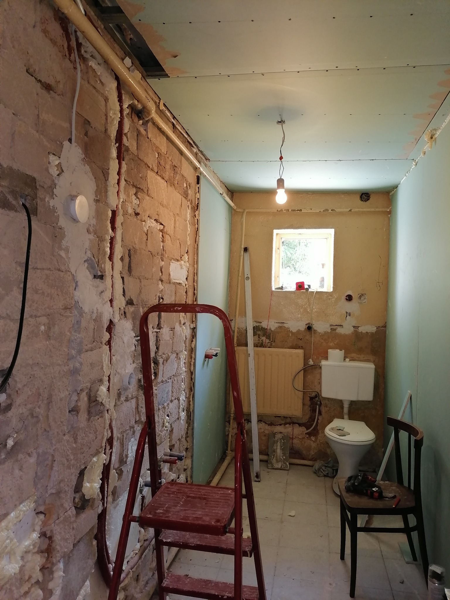Łazienka w trakcie remontu: odsłonięta ceglana ściana z pianką montażową, częściowo położona płyta gipsowa na ścianach i suficie, drabina malarska, toaleta, krzesło z wiertarką, grzejnik i okno...