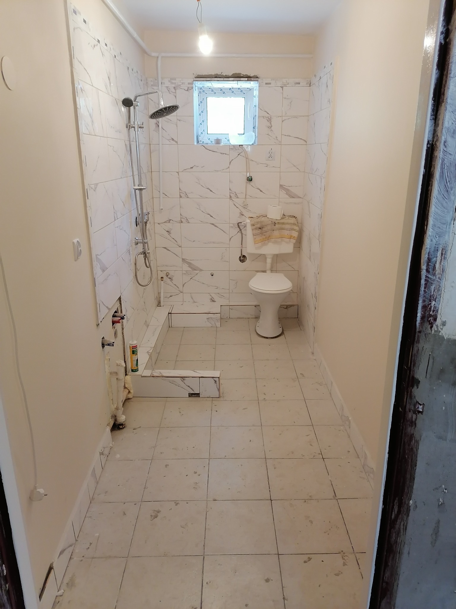 Łazienka w trakcie prac wykończeniowych: białe płytki imitujące marmur na ścianach, częściowo ułożone, widoczny brodzik prysznicowy z zamontowaną armaturą, toaleta z założonym ręcznikiem, podłoga...