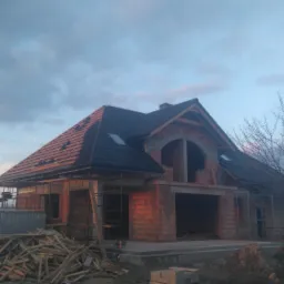 Surowy stan dwukondygnacyjnego domu z czerwonej cegły z częściowo ukończonym dachem z czarnej dachówki, widoczne rusztowania i stos drewna na pierwszym planie, pochmurne niebo w tle.