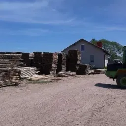 Stosy tarcicy różnej grubości i szerokości na placu, w tle budynek mieszkalny i operator w zielonej ładowarce kołowej.