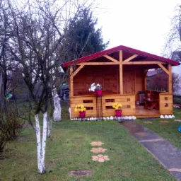 Drewniana altana z czerwonym dachem, widoczna z zewnątrz, z drewnianą ławeczką w środku, ozdobiona kwiatami w różowych doniczkach, z drzewem o bielonych pniach na pierwszym planie.