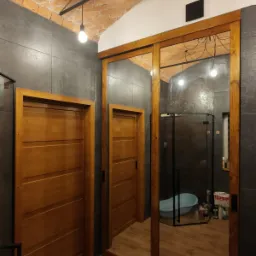 Nowoczesna łazienka z przesuwnymi drzwiami z drewnianą ramą, odbijającymi wnętrze z kabiną prysznicową i wanną na tle szarych płytek i ceglanego sufitu.