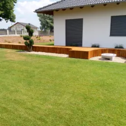 Drewniana platforma tarasowa z siedziskami, obłożona drobnym kamieniem, przylegająca do białego domu z szarymi roletami, na tle zielonego trawnika.