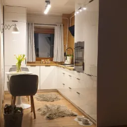 Biała kuchnia na wymiar z drewnianym blatem i podłogą, okrągły stół z bukietem tulipanów, dwa dywaniki imitujące skórę owczą, miski dla zwierząt przy ścianie.