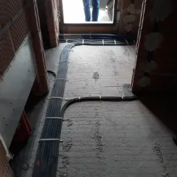 Kable elektryczne w peszlach ułożone na podłodze w trakcie prac remontowych w Tarnowie, widok z perspektywy niskiego kąta. W tle widoczne fragmenty muru i osoba w drzwiach.