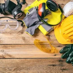 Komplet środków ochrony osobistej BHP: żółty kask, okulary ochronne, maska przeciwpyłowa, rękawice robocze i nauszniki wygłuszające, ułożone na drewnianym blacie.