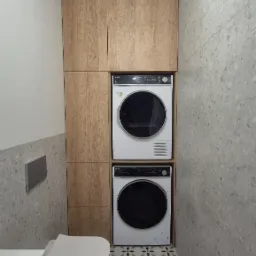 Mała łazienka z zabudową meblową z drewna, w której umieszczono pralkę i suszarkę jedna na drugiej, wisząca toaleta, ściany wykończone płytkami imitującymi lastryko, podłoga z wzorem kwiatowym.