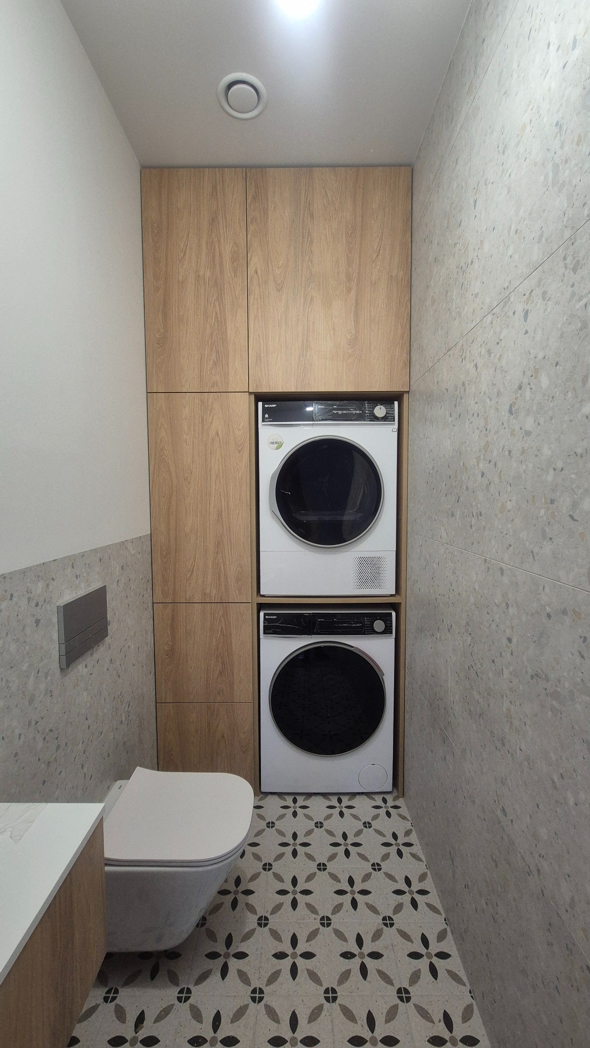 Mała łazienka z zabudową meblową z drewna, w której umieszczono pralkę i suszarkę jedna na drugiej, wisząca toaleta, ściany wykończone płytkami imitującymi lastryko, podłoga z wzorem kwiatowym.