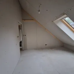 Pokój na poddaszu w trakcie remontu, ściany i skosy wyłożone płytami karton gips, widoczne drewniane elementy konstrukcyjne, okno dachowe i wisząca żarówka.