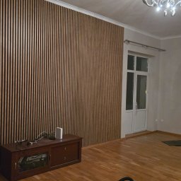 USŁUGI BUDOWLANO SPRZĄTAJĄCE R&B - Ściana wykończona panelami lamelowymi w odcieniach brązu, tworzącymi pionowy wzór. Widoczny fragment pokoju z parkietem, szafką RTV i drzwiami balkonowymi.