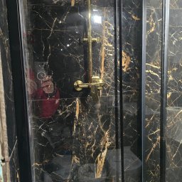 USŁUGI BUDOWLANO SPRZĄTAJĄCE R&B - Kabina prysznicowa z czarnymi płytkami imitującymi marmur ze złotymi żyłami i złotą armaturą. Widoczne odbicie osoby robiącej zdjęcie.